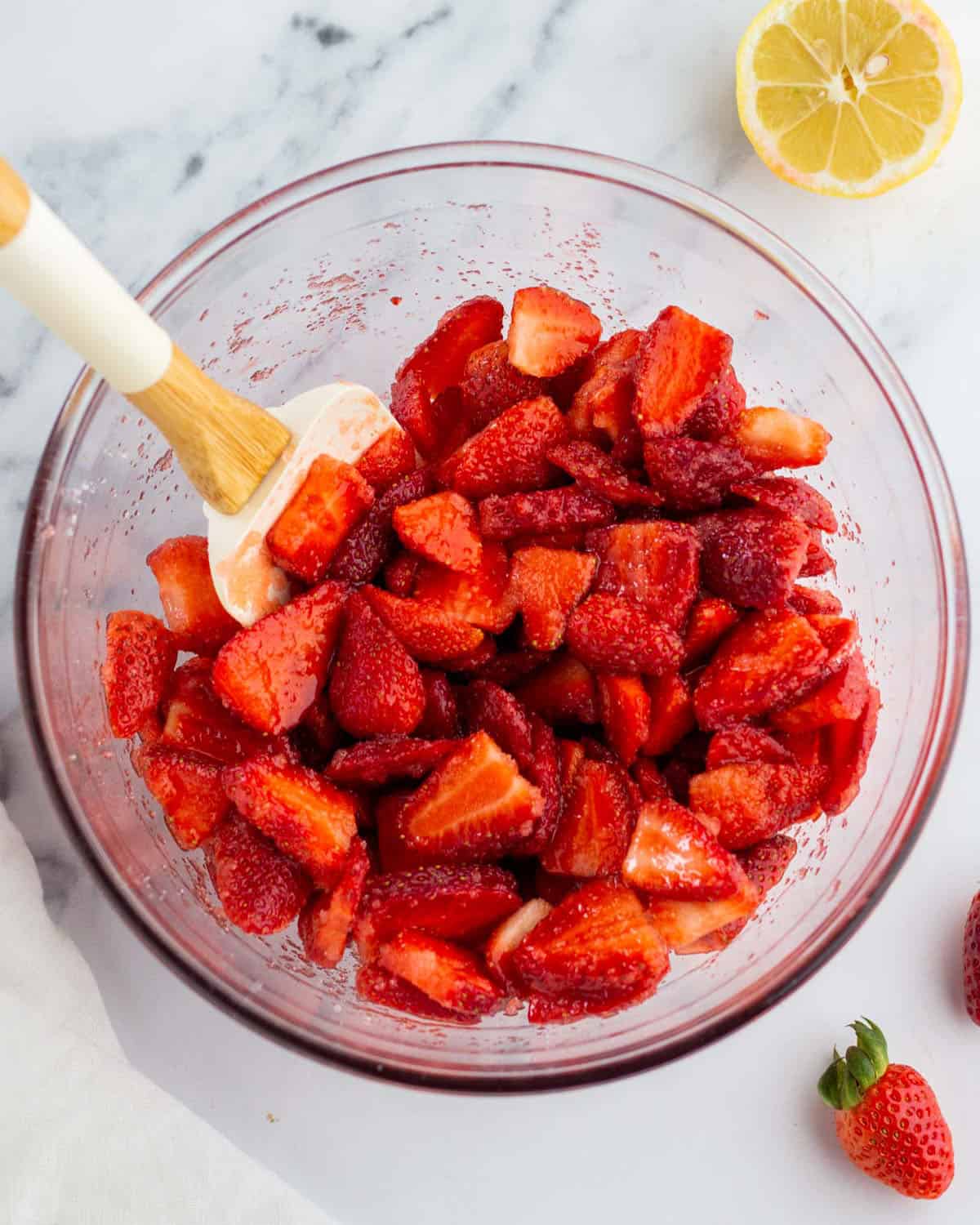 A clear mixing bowl with sliced strawberries being stirred with sugar, surrounded by a fresh lemon half and fresh whole strawberries. 
