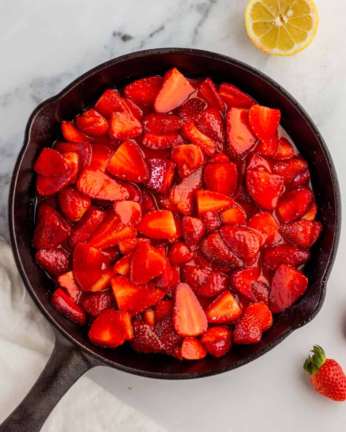 Sugared strawberries in a layer in a cast iron skillet, surrounded by fresh strawberries and lemon. 