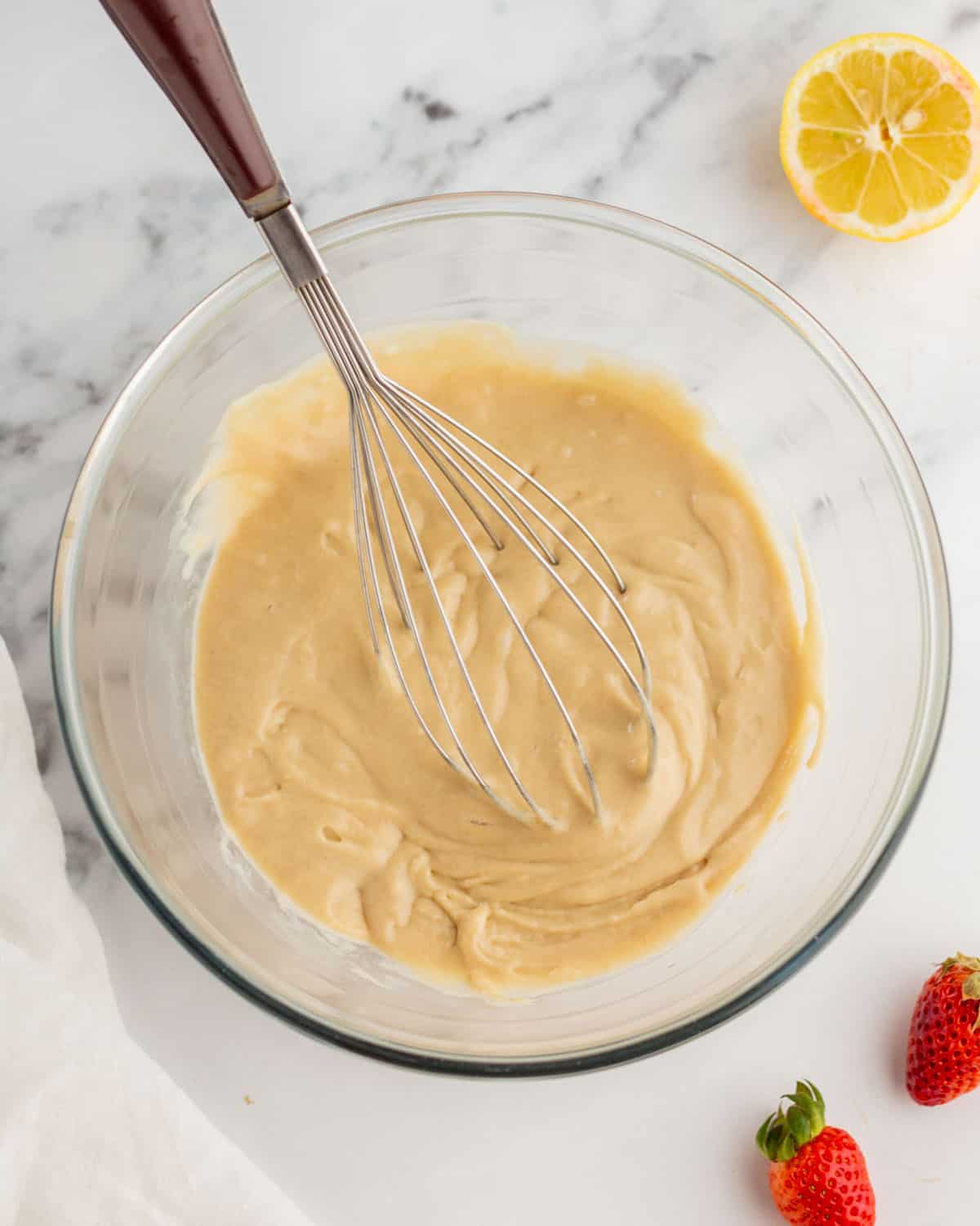 Batter in a bowl with a whisk surrounded by fresh strawberries and a lemon. 