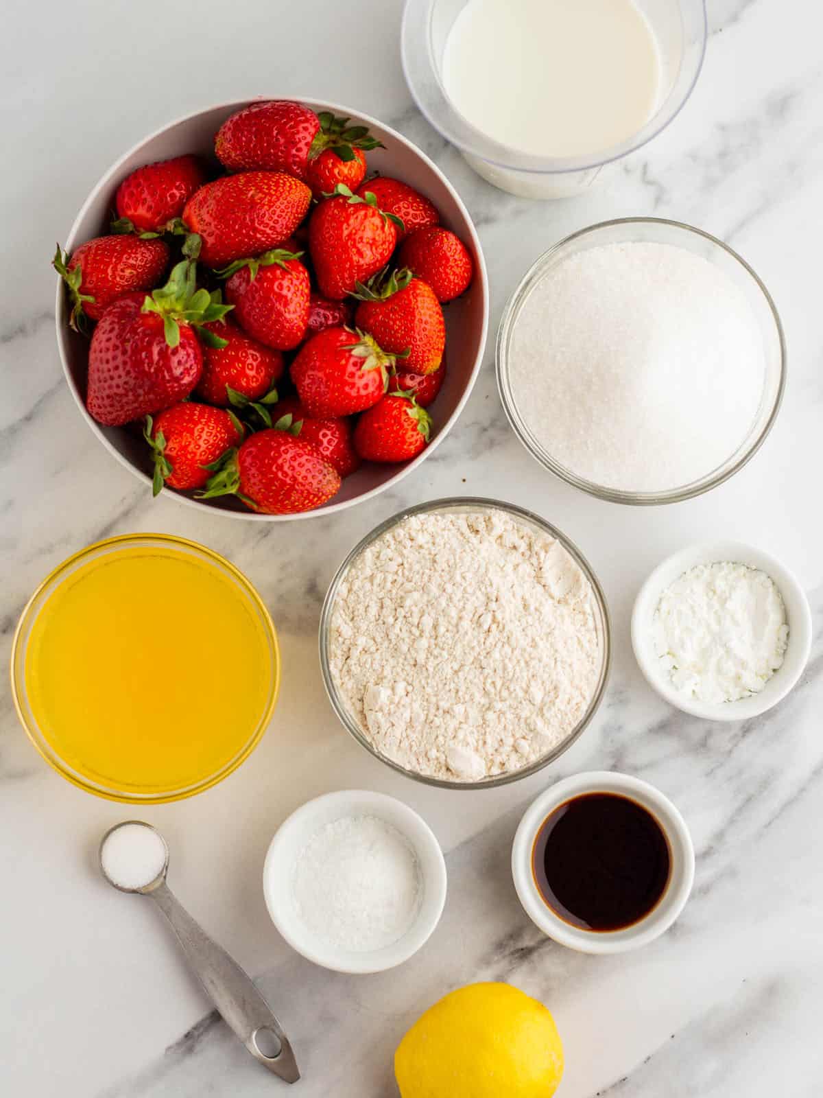 Ingredients in bowls on a white and gray granite counter. Fresh strawberries, flour, baking powder, lemon juice, and more. Surrounded by a fresh lemon and a measuring spoon. 
