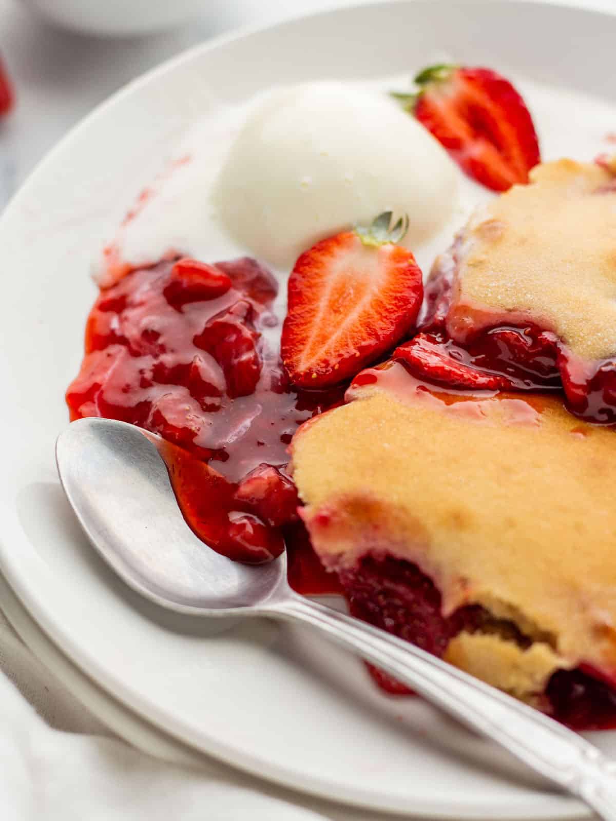 A plate with warm strawberry cobbler with a spoon and a scoop of vanilla ice cream and a fresh strawberry. 