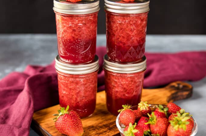 Two stacks of two jars of canned strawberry jam, with fresh strawberries on top, placed on a wooden cutting board. A bowl of fresh strawberries and a burgundy cloth surrounds.