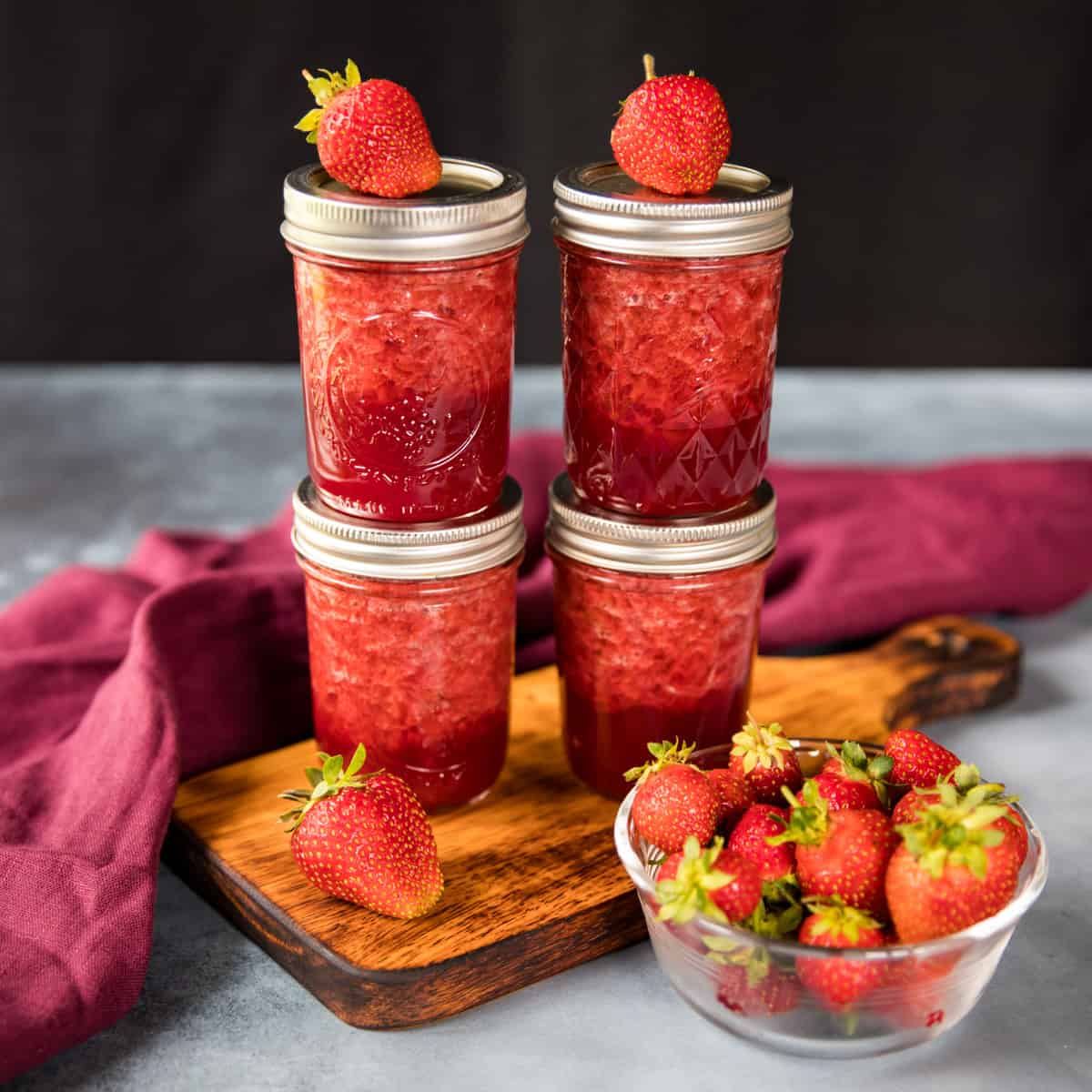 Two stacks of two jars of canned strawberry jam, with fresh strawberries on top, placed on a wooden cutting board. A bowl of fresh strawberries and a burgundy cloth surrounds.