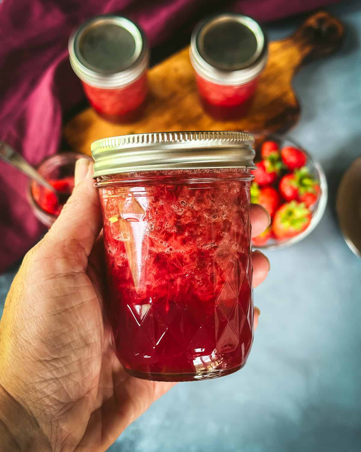 A hand holding a jar of strawberry jam with more jars and fresh strawberries in the background.