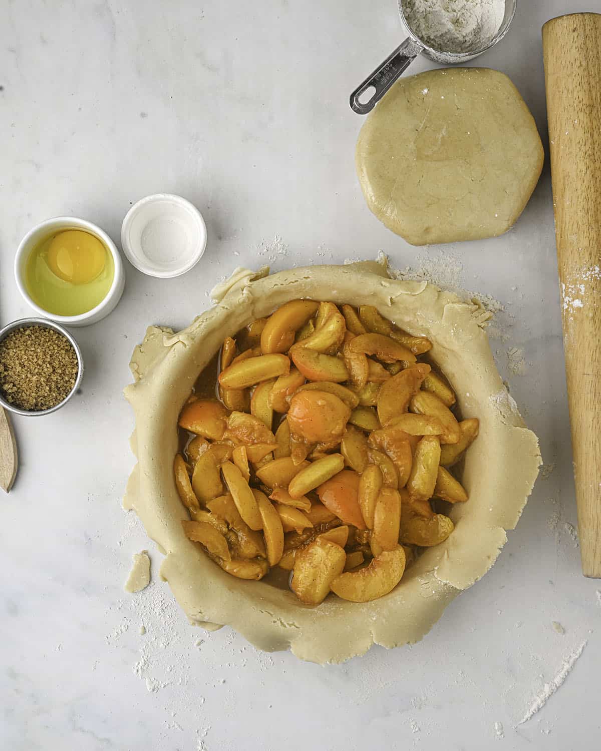 A pie crust pan with pie dough and peach pie filling in it, with other peach pie ingredients in bowls surrounding. 
