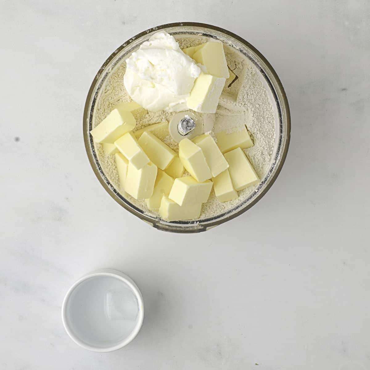 Dry ingredients topped with butter in a food processor, top view. 