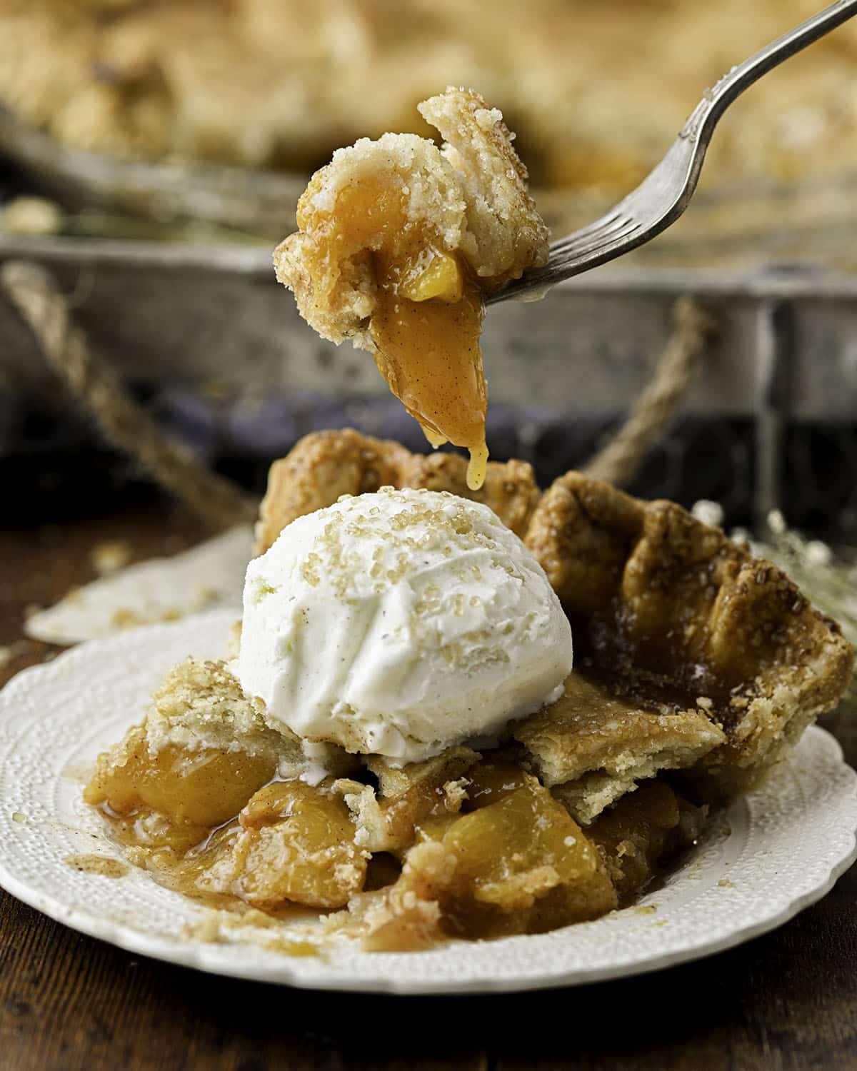 A slice of peach pie on a white plate topped with vanilla ice cream, with a fork holding up a bite of the pie. 