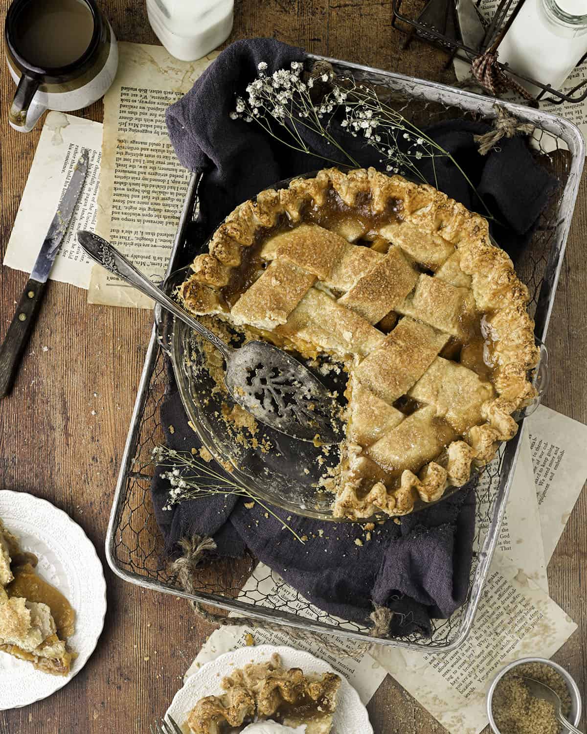 Top view of a cut peach pie in a basket with a dark cloth, flowers, and surrounded by plates of pie and recipe pages. 
