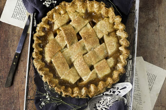 A peach pie, top view, in a wire basket with a dark towel underneath, with some baby's breath flowers and a pie server surrounding. All of a wood surface with recipe pages underneath the basket.