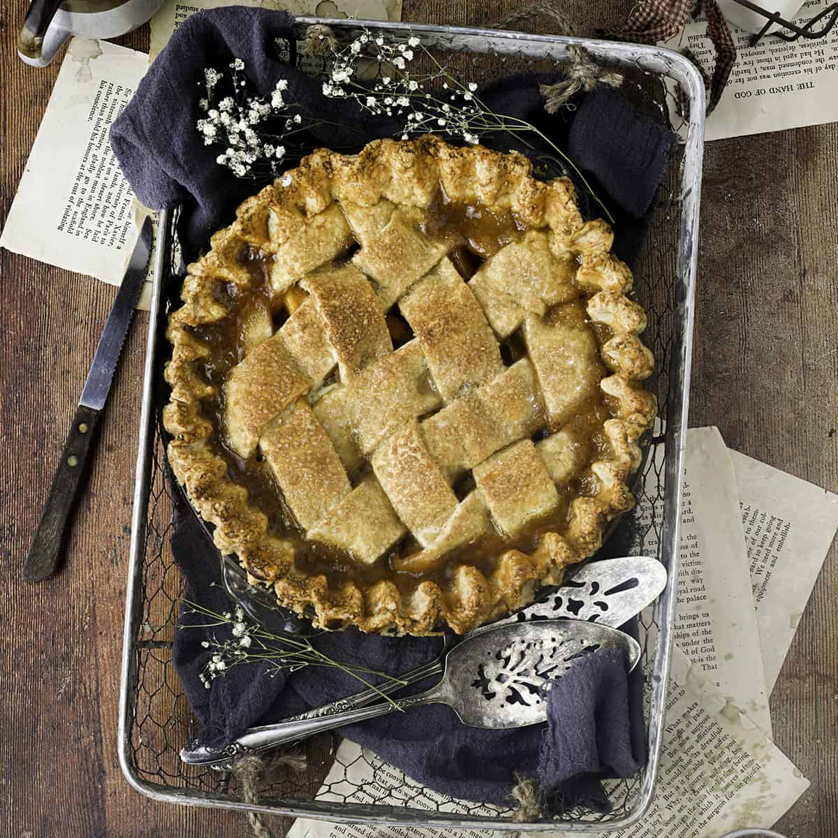 A peach pie, top view, in a wire basket with a dark towel underneath, with some baby's breath flowers and a pie server surrounding. All of a wood surface with recipe pages underneath the basket. 