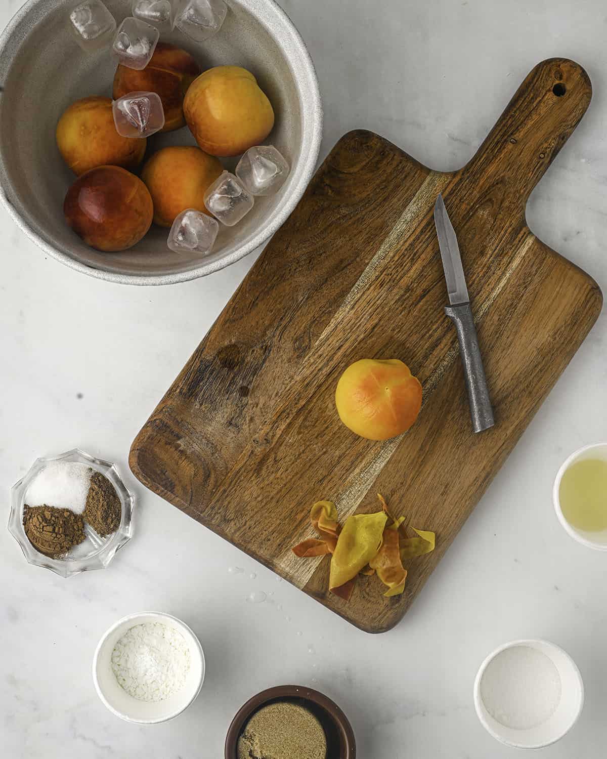 Blanched peaches in a bowl of ice, and one on a wood cutting board that is ready to be sliced, with small bowls of peach filling ingredients surrounding. 