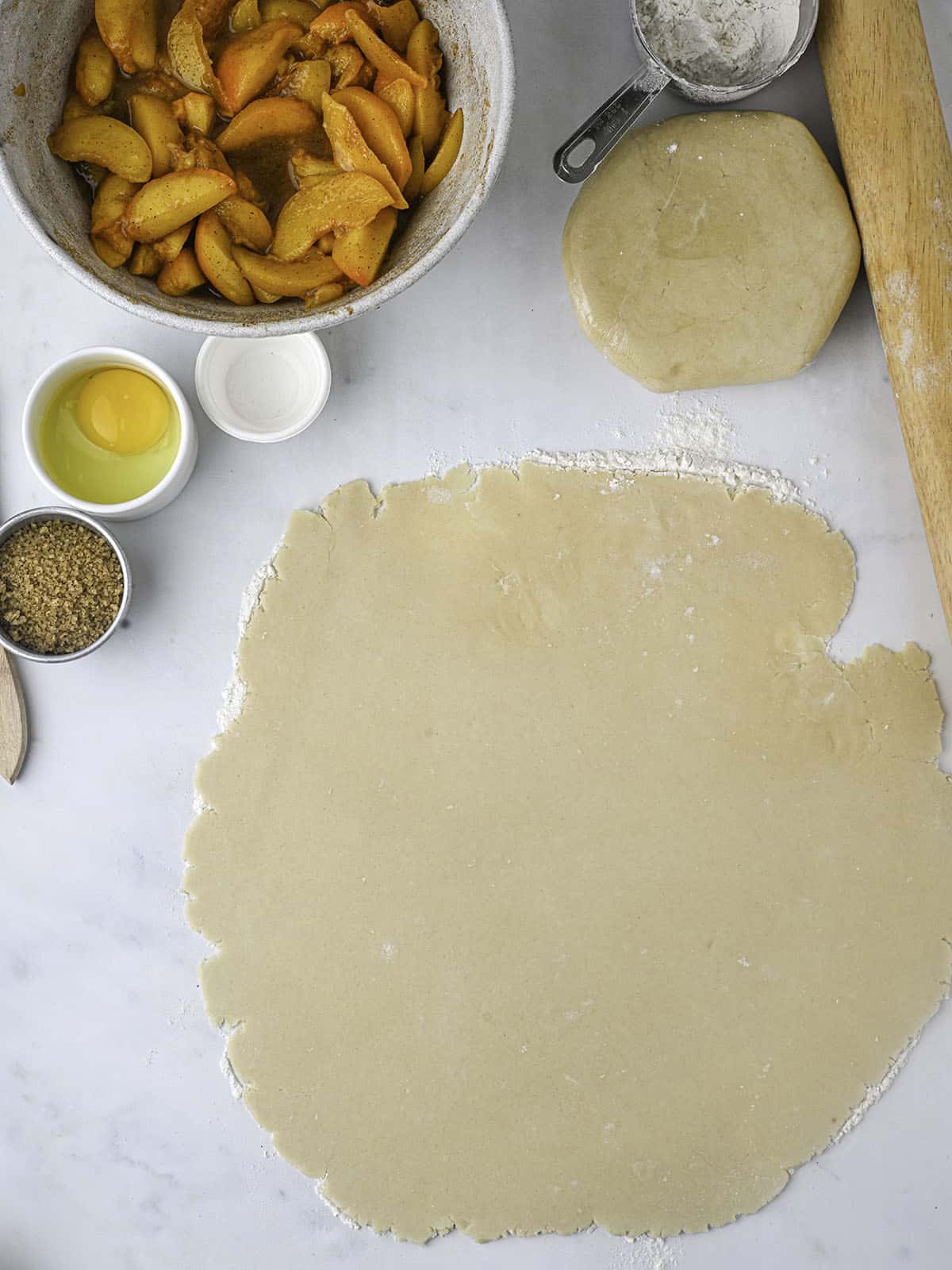 A pie crust rolled out into a rough circle shape, surrounded by peach pie filling in a bowl and other ingredients in small bowls. 