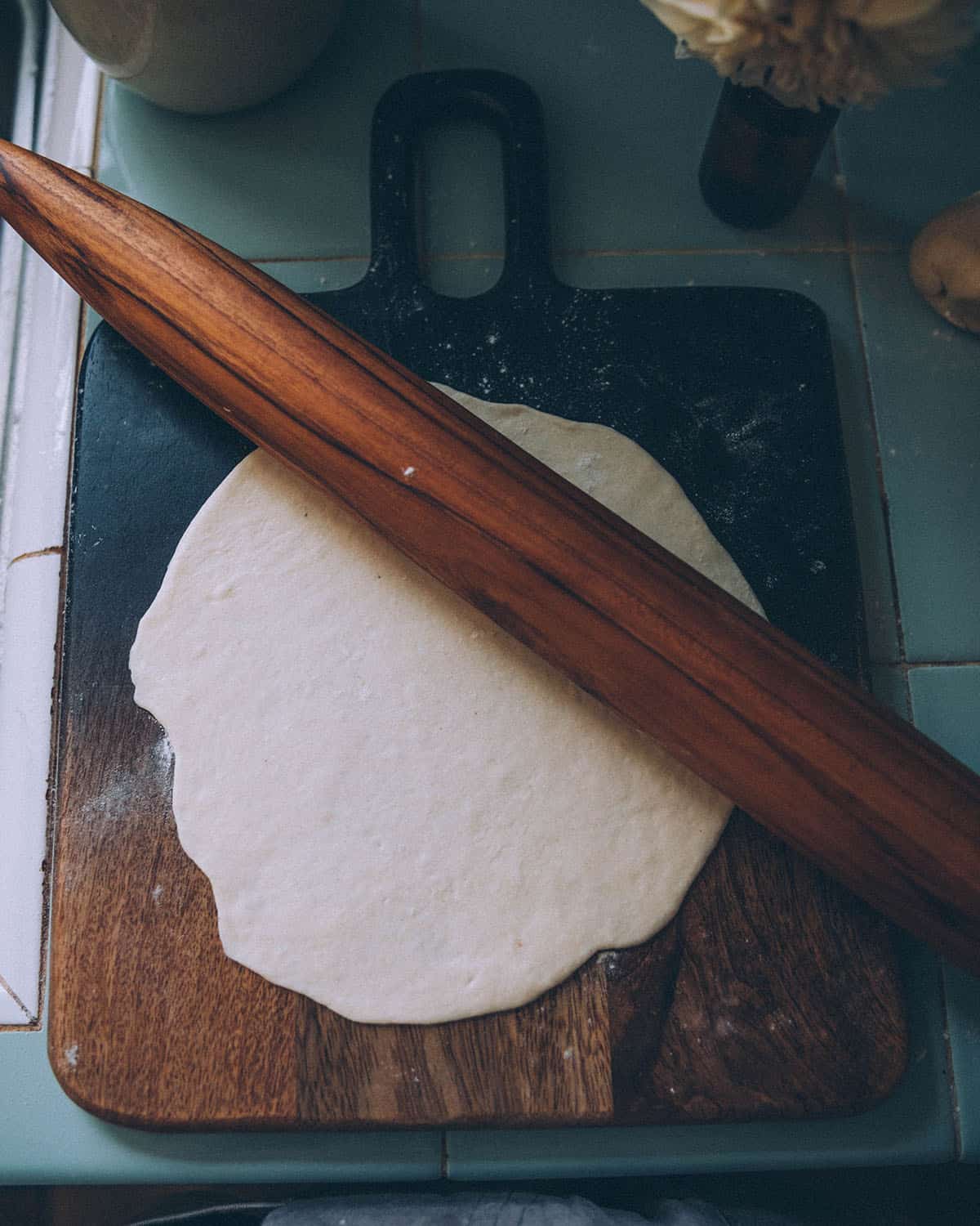 A dark wood cutting board with a round dough roll out with a dough roller set on top. 