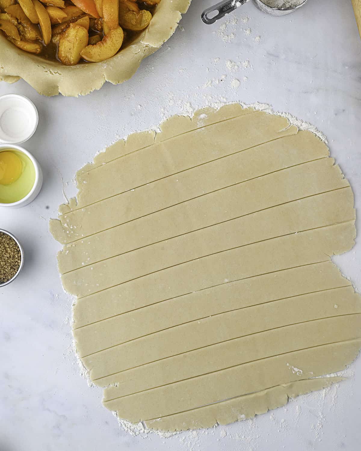 A circle of pie dough rolled out and cut into strips with other peach pie ingredients surrounding. 