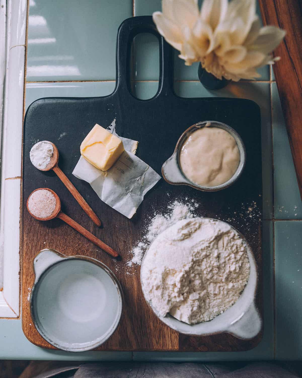 A dark wood cutting board on a mint tile surface, with bowls of ingredients for sourdough tortillas. Top view. 