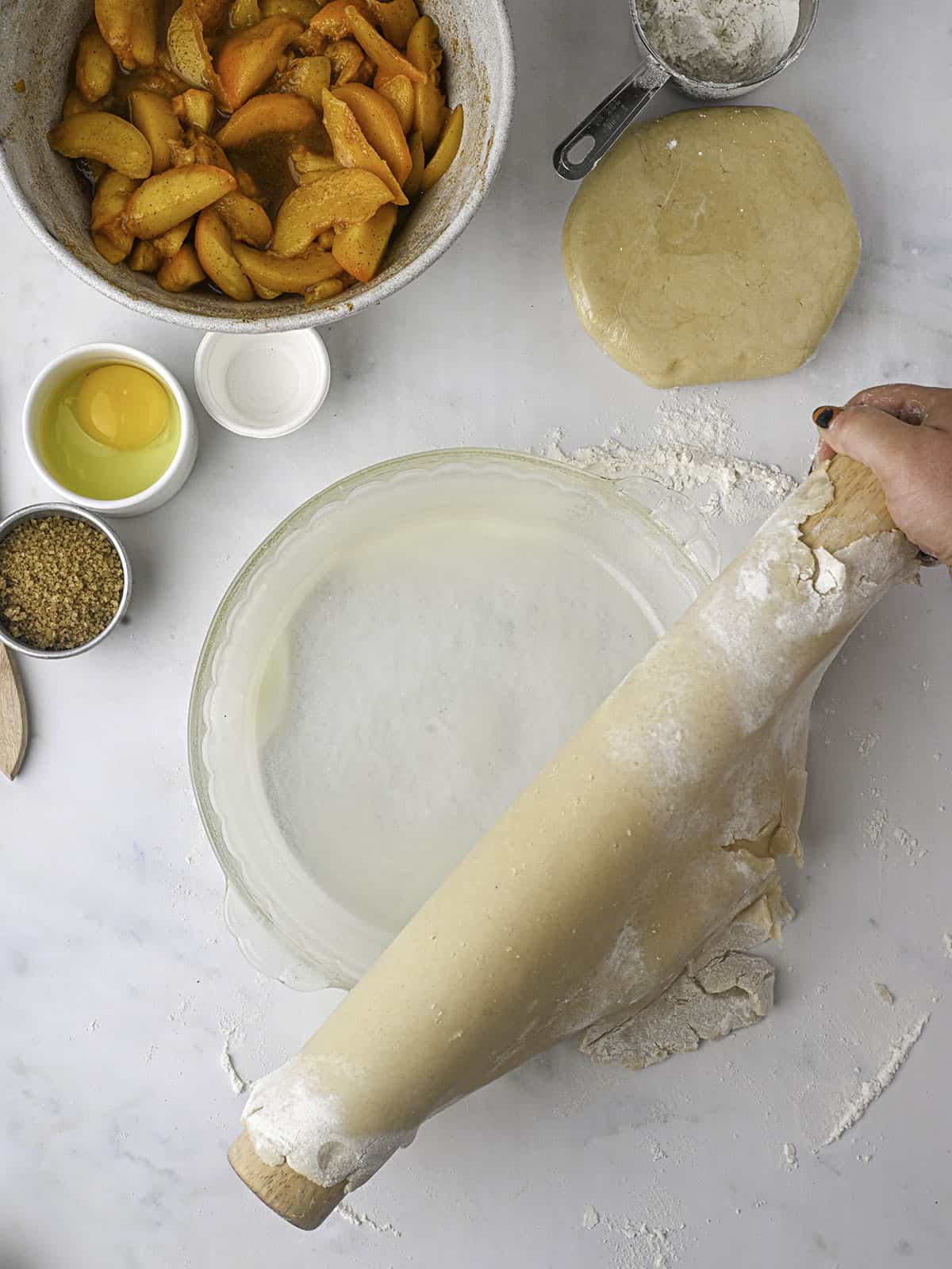 Pie dough transferring to a pie pan with a rolling pin, with peach pie ingredients in bowls surrounding. 