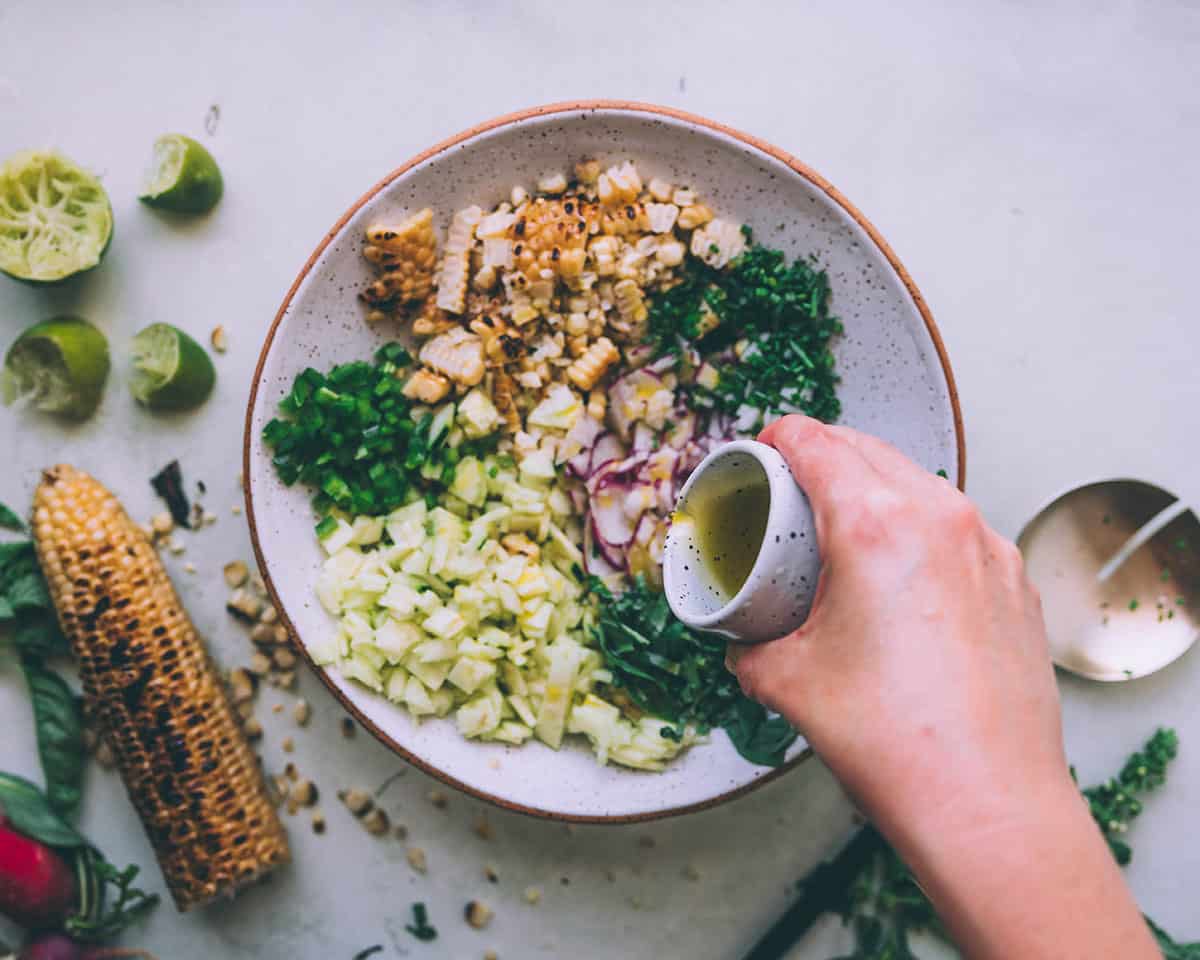 Olive oil pouring from a small bowl into a larger bowl of corn salad.