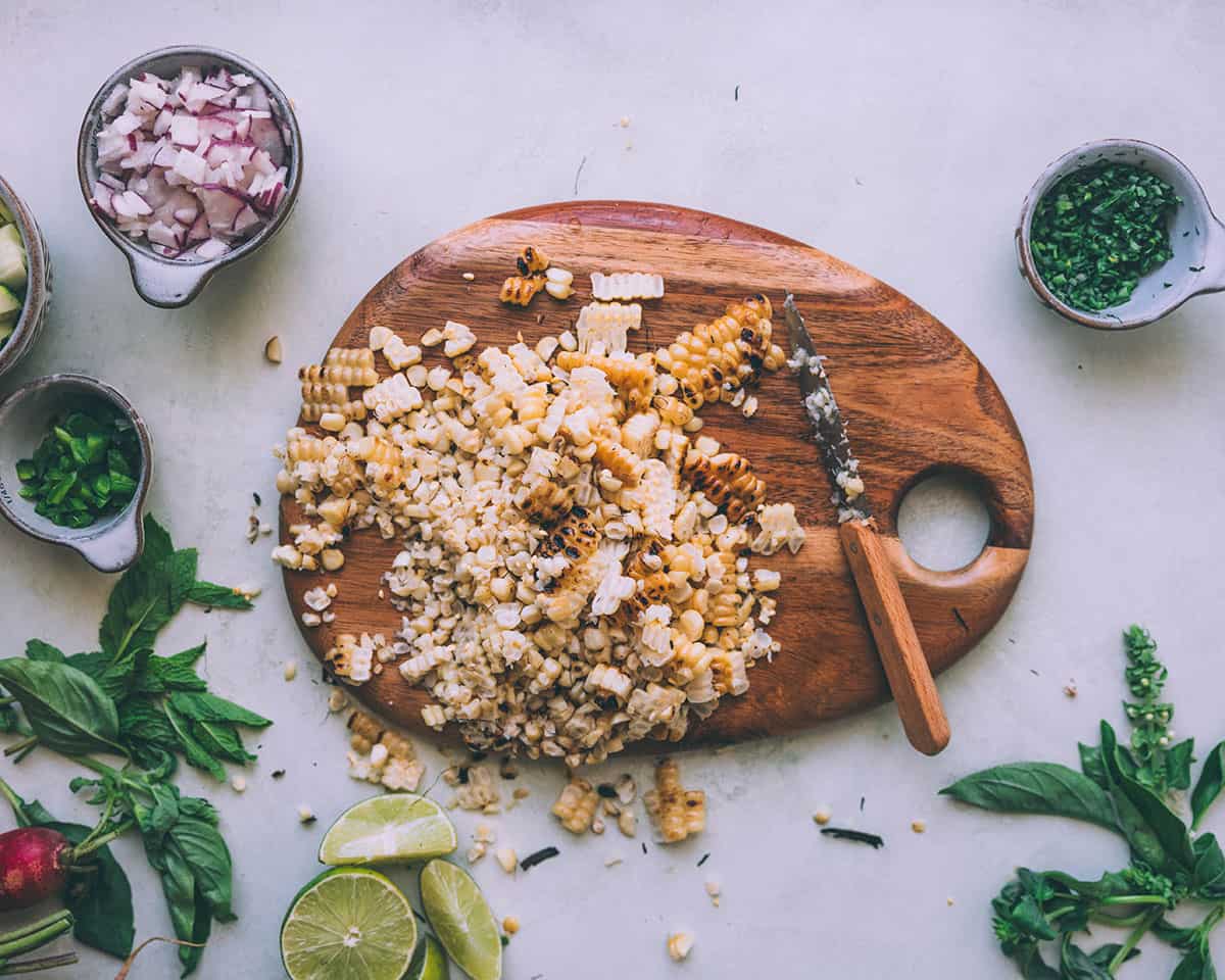 Corn chopped off the cob on a wood cutting board surrounded by other salad ingredients. 