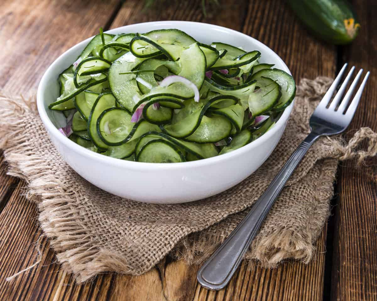 A white bowl filled with sliced cucumber salad on a wood surface with a burlap placemat and a fork laying to the side. 