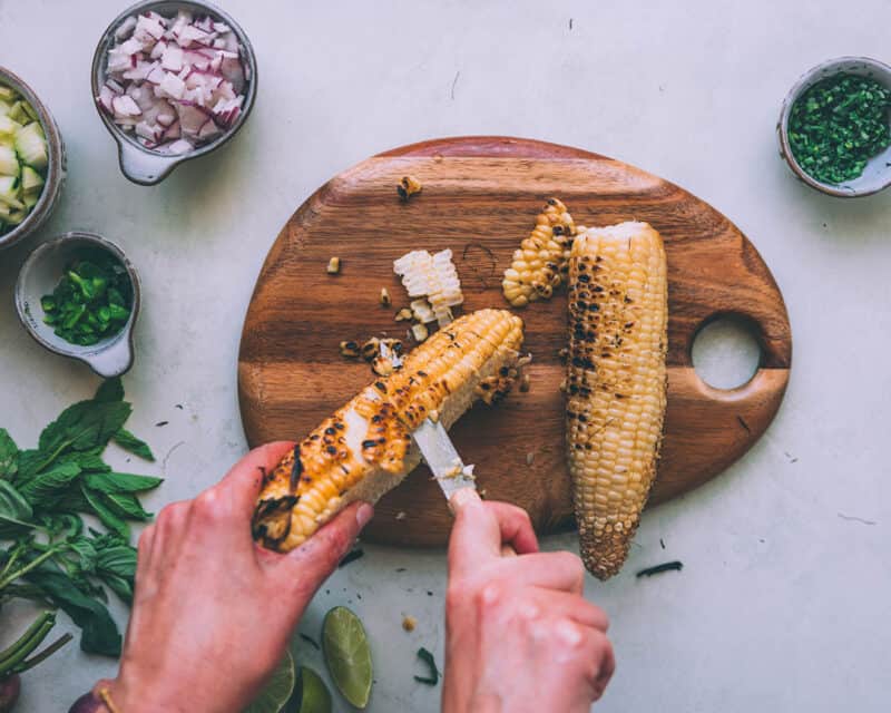 A wooden cutting board with corn being cut off the cob with a knife. 
