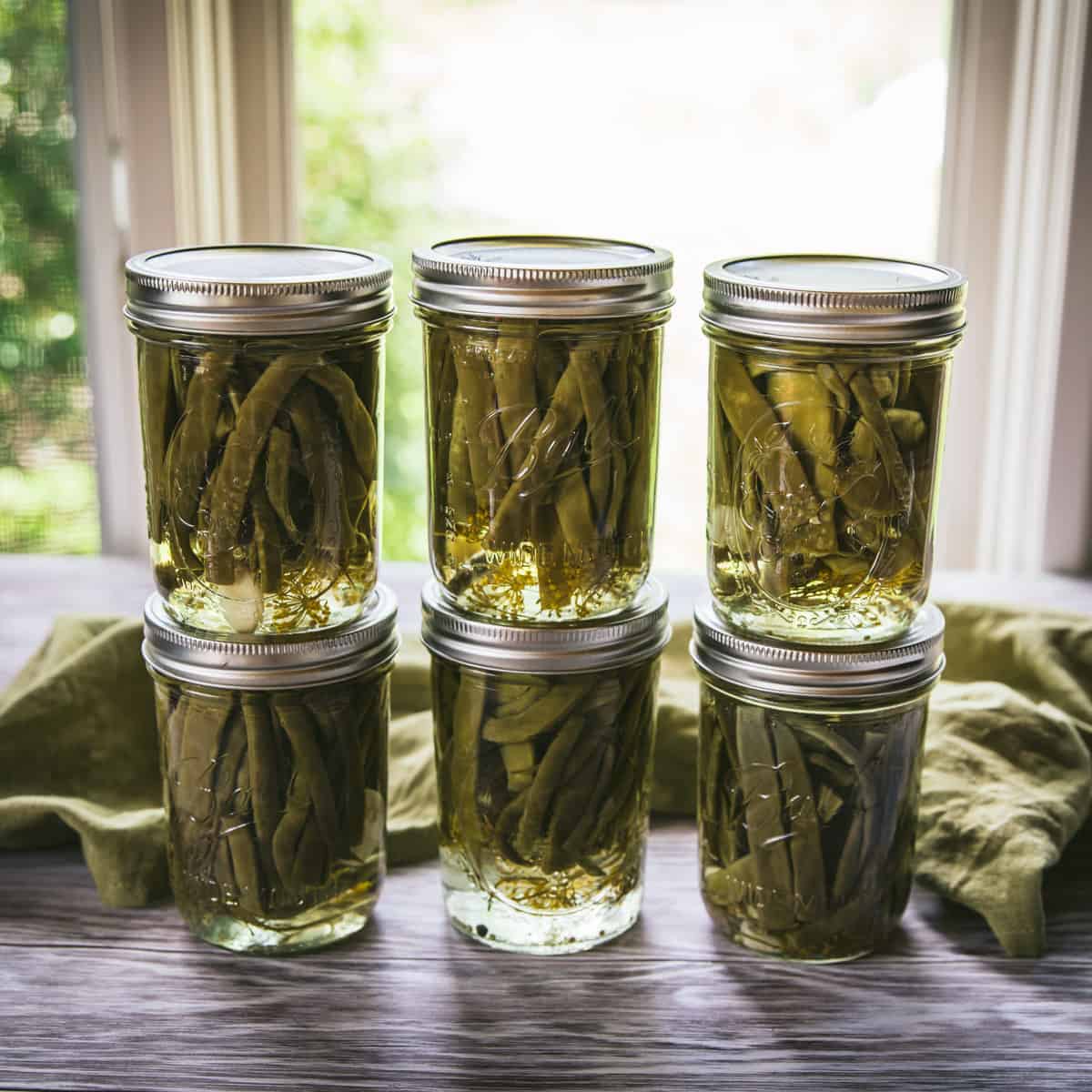 6 cans of dilly beans stacked in 2's on a counter with a window in the background. 