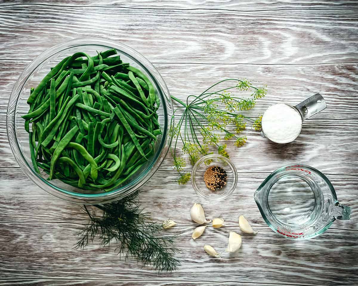 Green beans in a bowl on a gray wood surface, surrounded by a dill flower, pickling salt, and other dilly bean ingredients. 