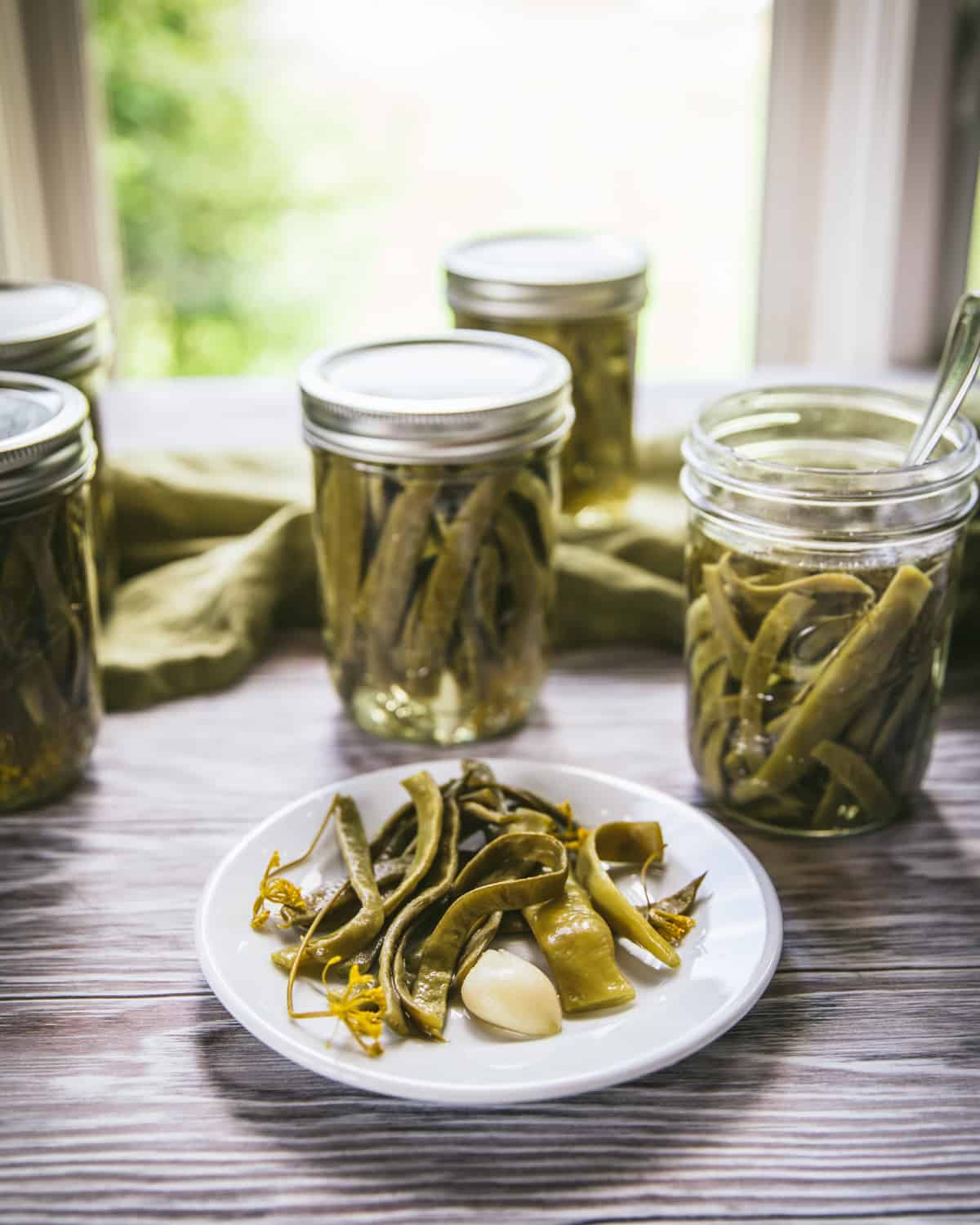 Dilly beans on a white plate on a wood surface with jars in the background, side view.