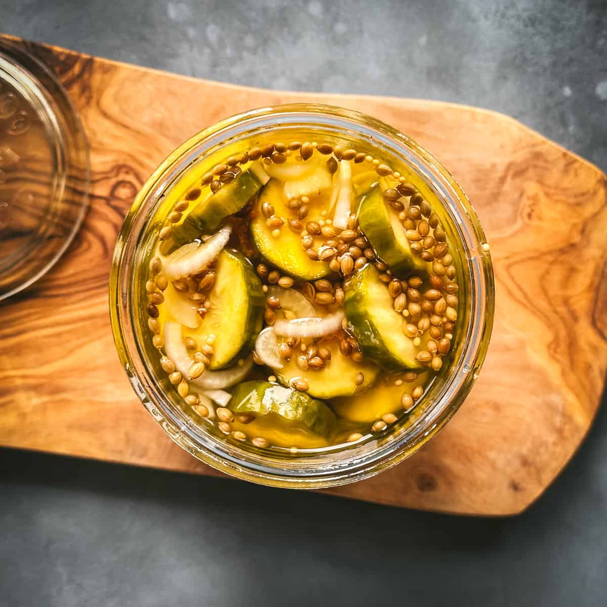 Top view of jar of bread and butter pickles ready to eat, on a wood cutting board with a gray background. 