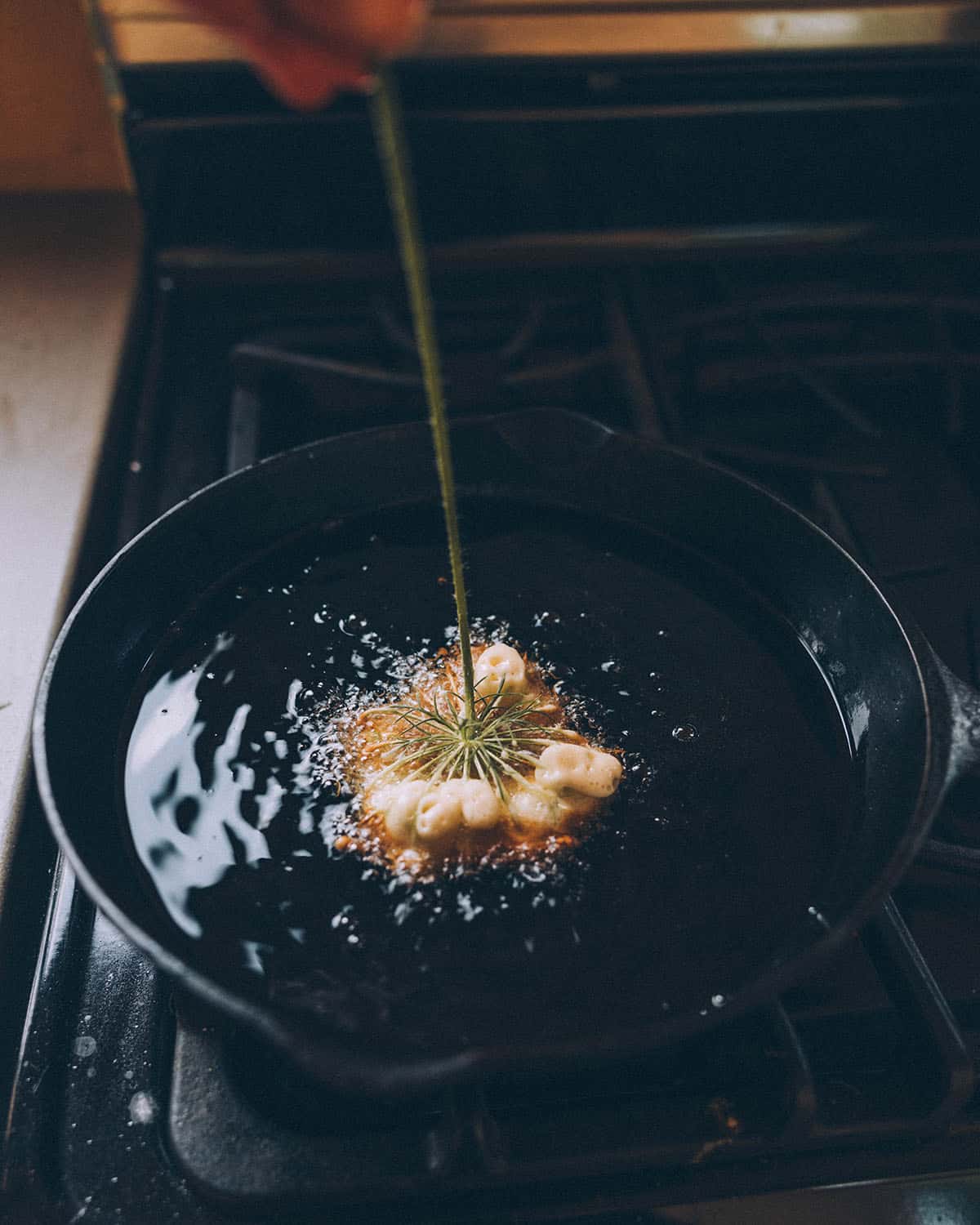 A Queen Anne's lace flower with batter, flower side down sizzling in hot oil in a black cast iron skillet. 
