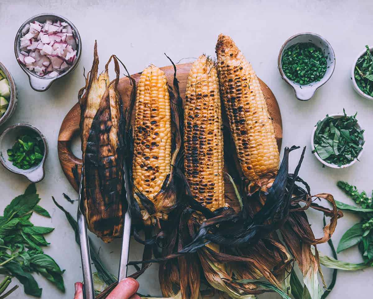 4 cobs of grilled corn on a wood cutting board, with bowls of salad ingredients and fresh herbs surrounding. 
