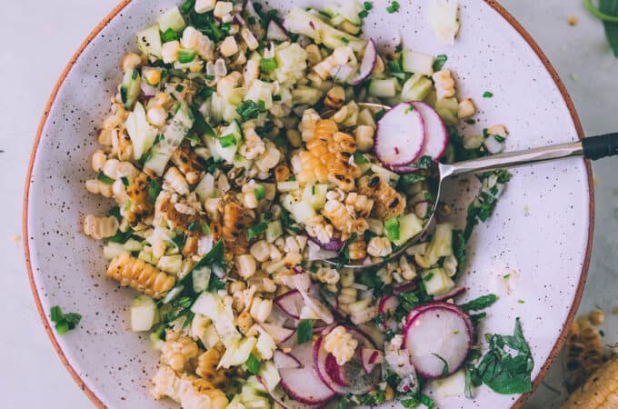 Grilled corn salad with radish and cucumber and fresh herbs, in a white bowl with a red rim, top view.