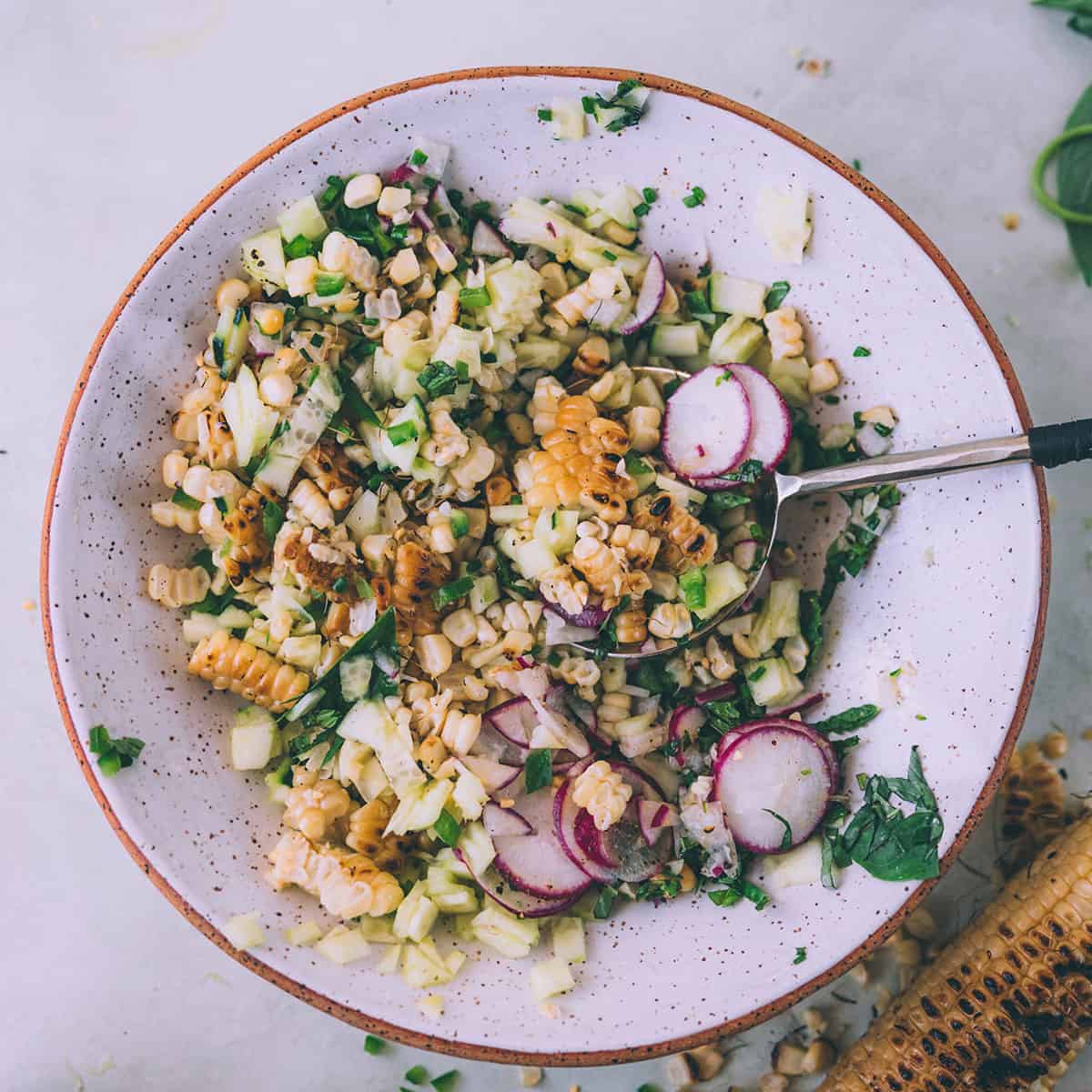 Grilled corn salad with radish and cucumber and fresh herbs, in a white bowl with a red rim, top view. 