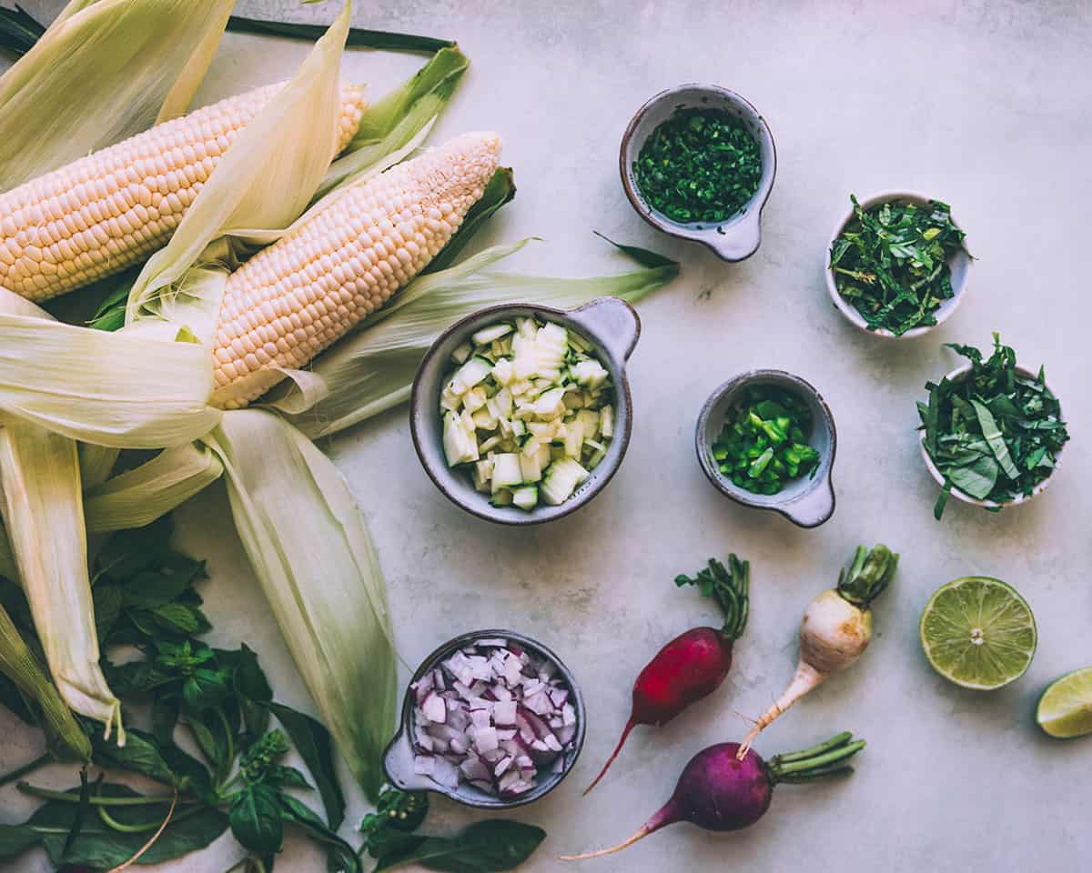 Corn salad ingredients on a white counter top, some chopped in bowls with fresh whole radishes, herbs, and corn surrounding. 