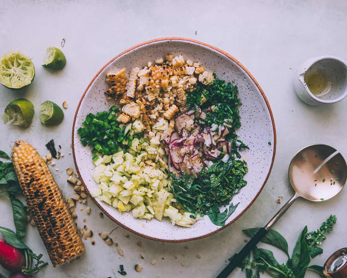 A bowl of all the corn salad ingredients, unmixed, surrounded by additional ingredients. 