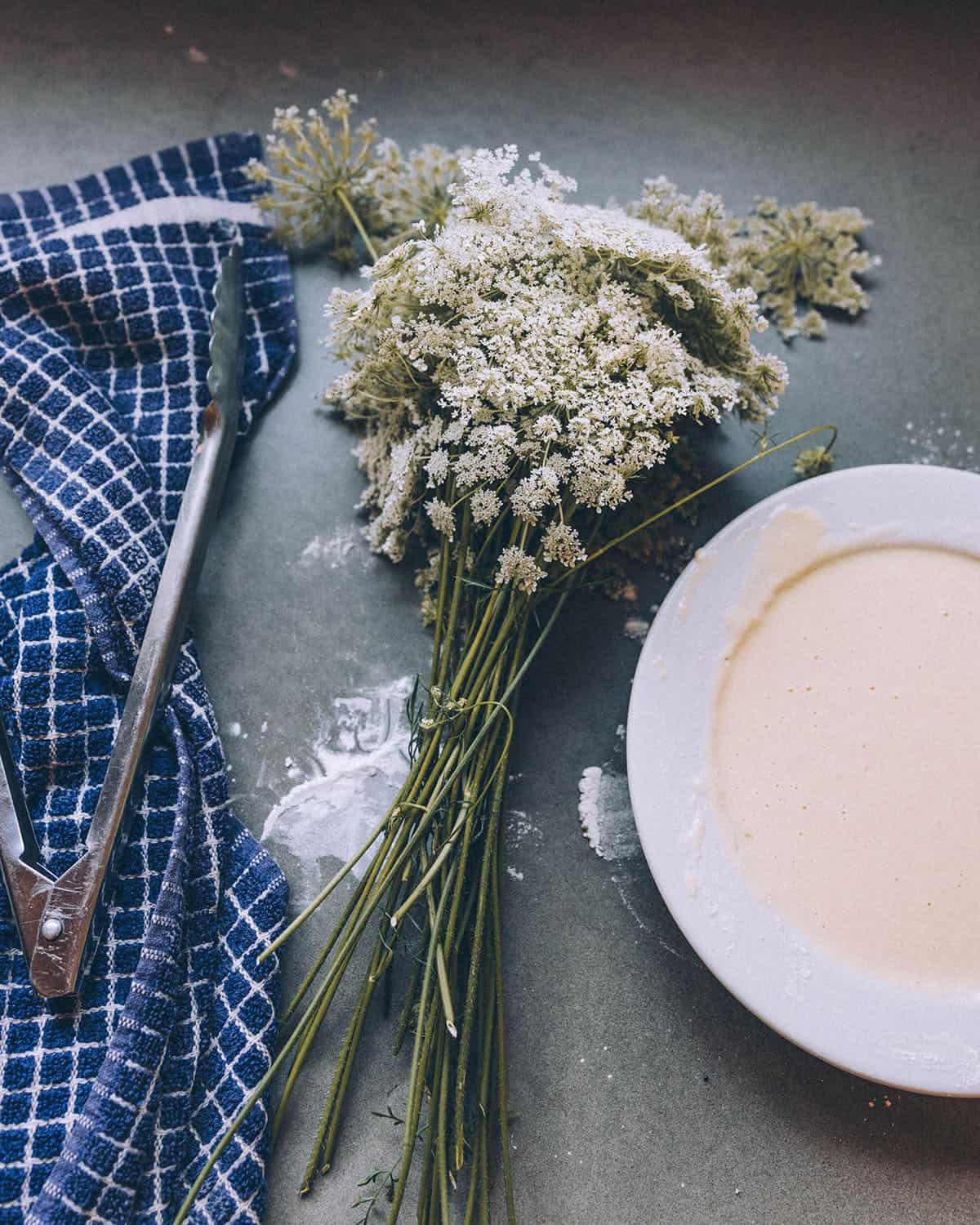 A white bowl of batter with fresh Queen Anne's lace flowers resting beside it with a towel and tongs.