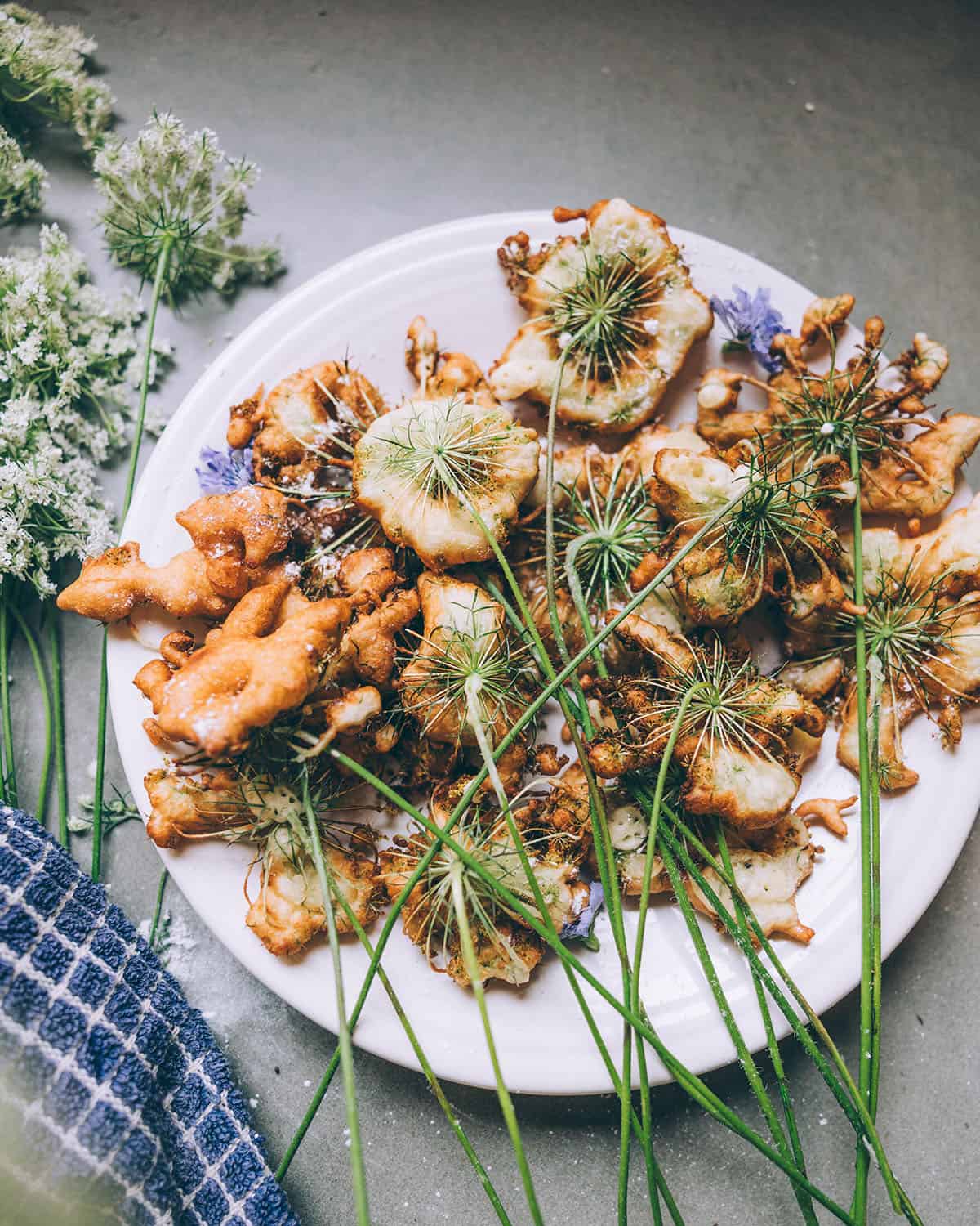 A white plate with Queen Anne's lace fritters stacked on it, all with green stems, on a gray counter surrounded by fresh Queen Anne's lace flowers. 