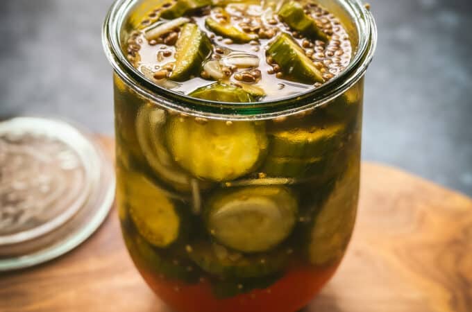 Bread and butter pickles in an open jar, on a wooden cutting board with a gray background.