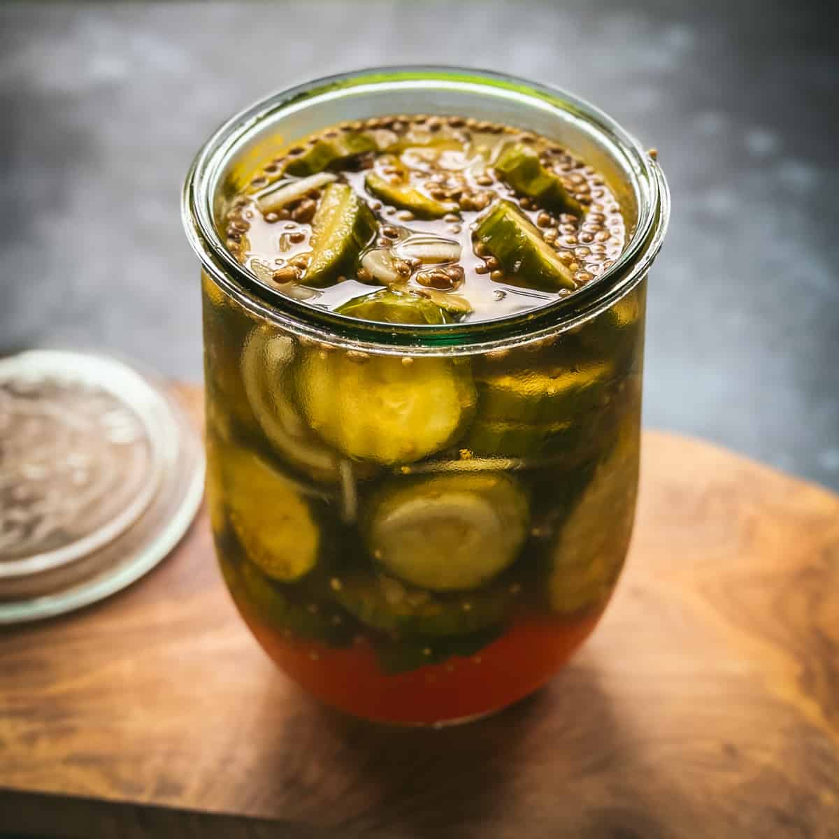 Bread and butter pickles in an open jar, on a wooden cutting board with a gray background. 