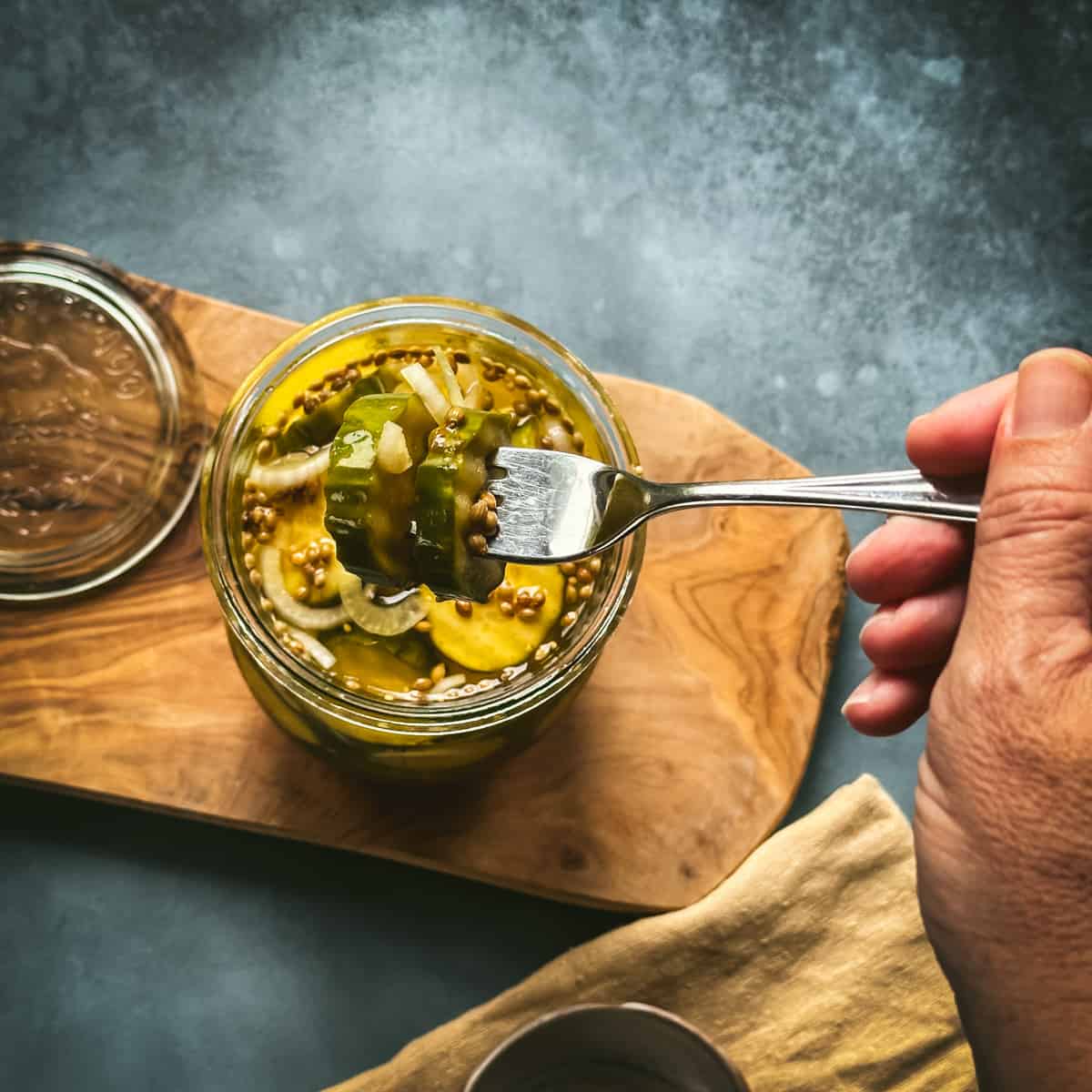 A jar of bread and butter pickles with a fork lifting some out of the jar, top view. Resting on a wood cutting board with a gray background. 