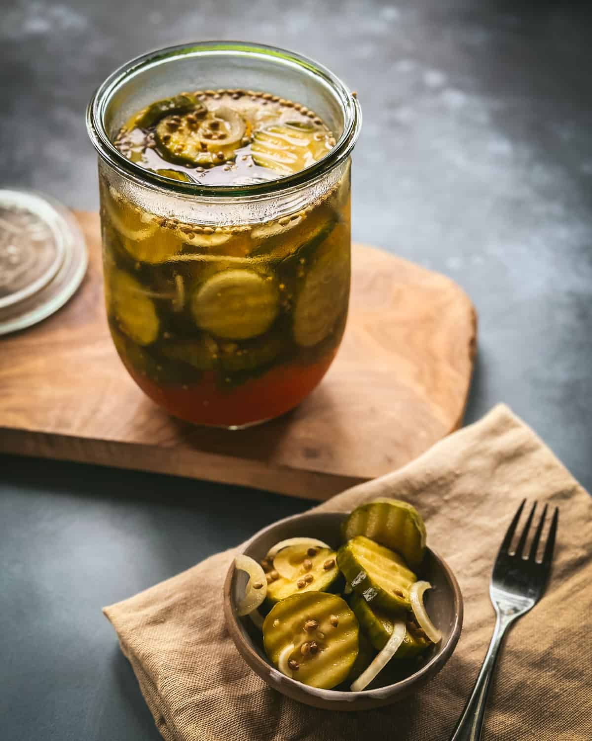 An open jar of pickles on a wood cutting board, with a small bowl of pickles next to it with a fork to the side, gray background. 