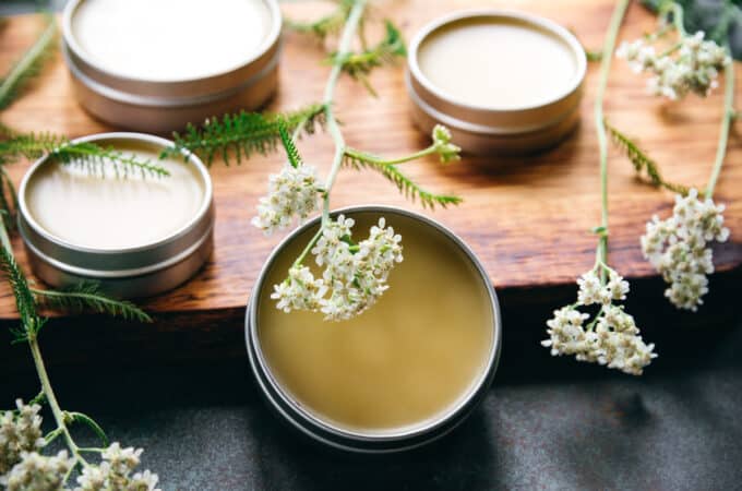 Yarrow salve in tins on a wooden cutting board, with yarrow flowers laying over the top, on a dark gray countertop.