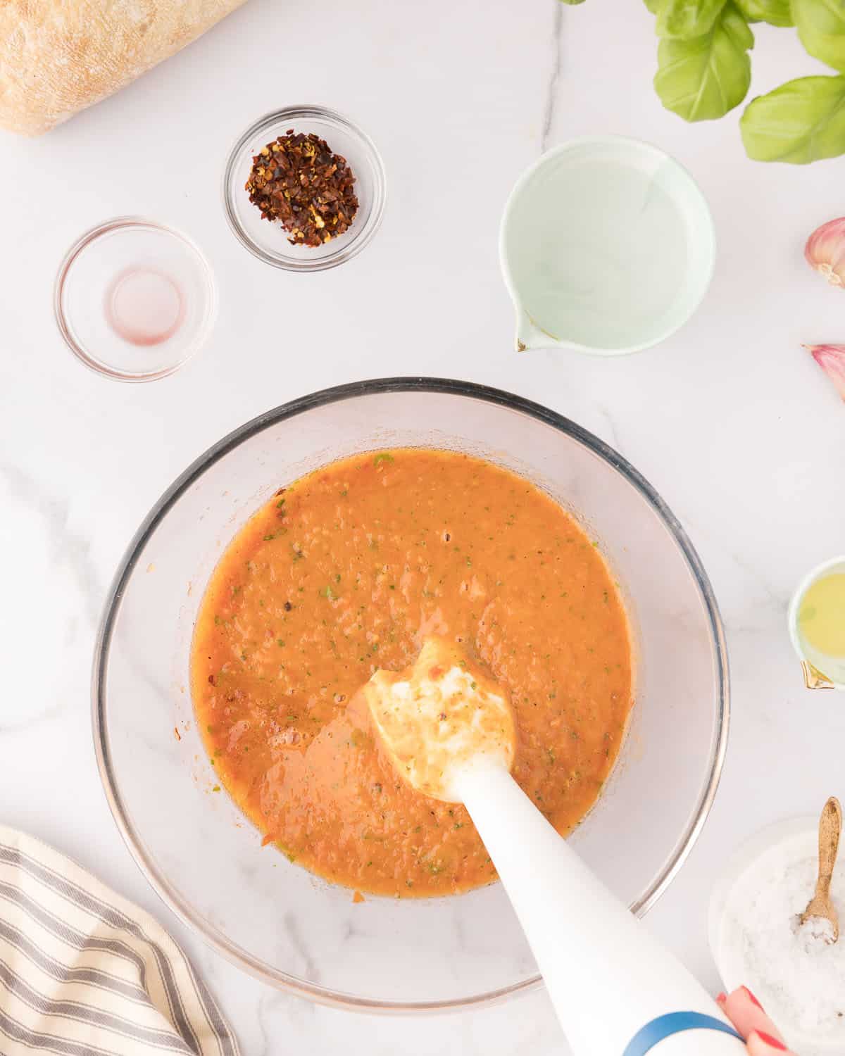 An immersion blender blending up the veggies for tomato soup in a clear bowl, with other ingredients in small bowls surrounding. 