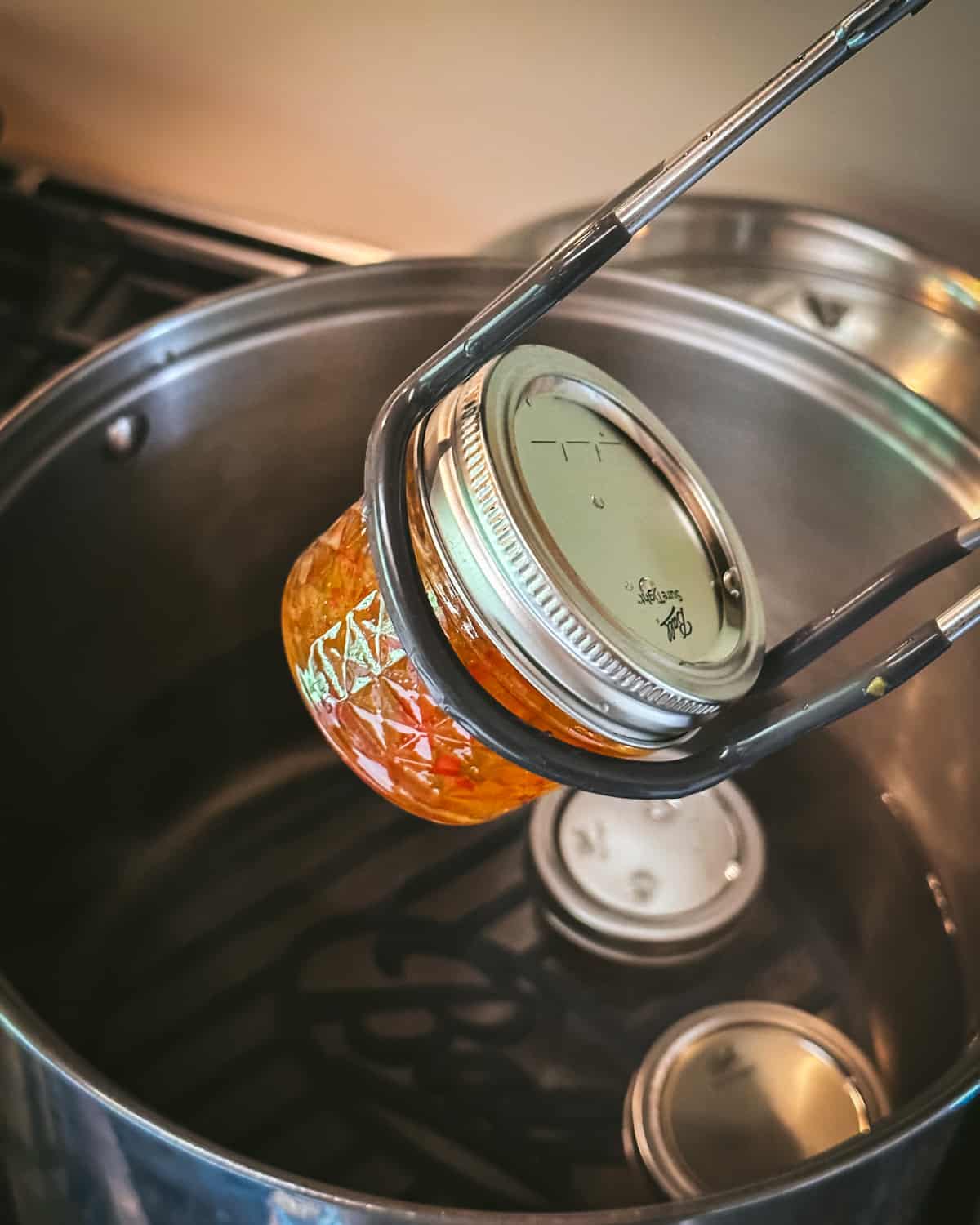 A small closed jar of pepper jelly being lifted out of a hot water bath canner with a jar lifter. 