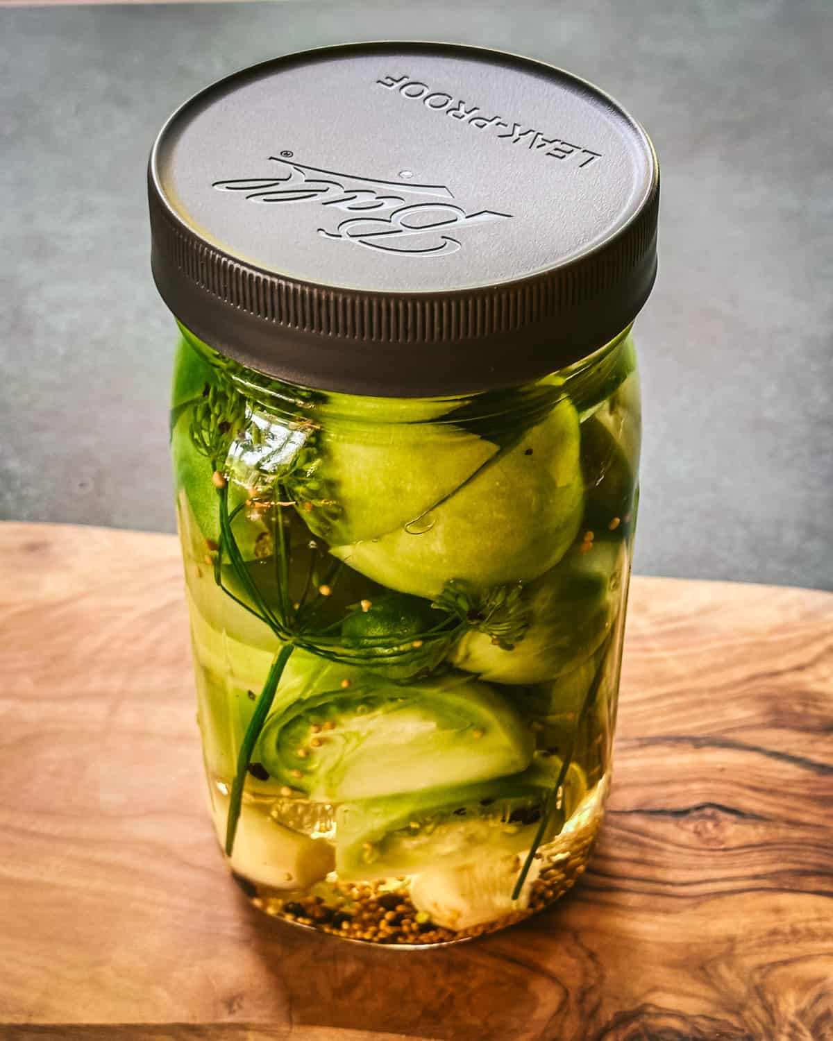 Green tomato wedges in brine, in a jar with a lid on a wood cutting board. 