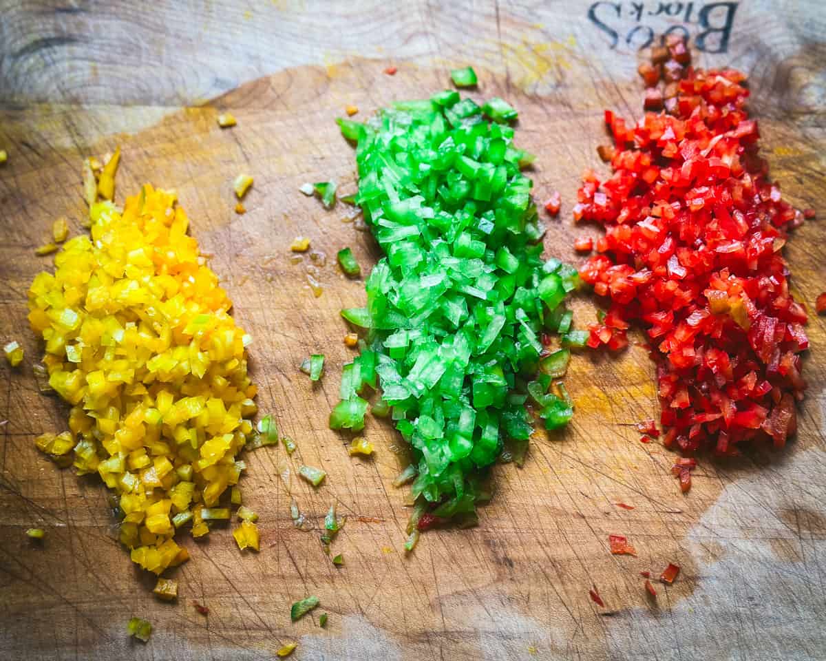 A wooden cutting board with 3 piles of chopped bell peppers, yellow, green, and red. 