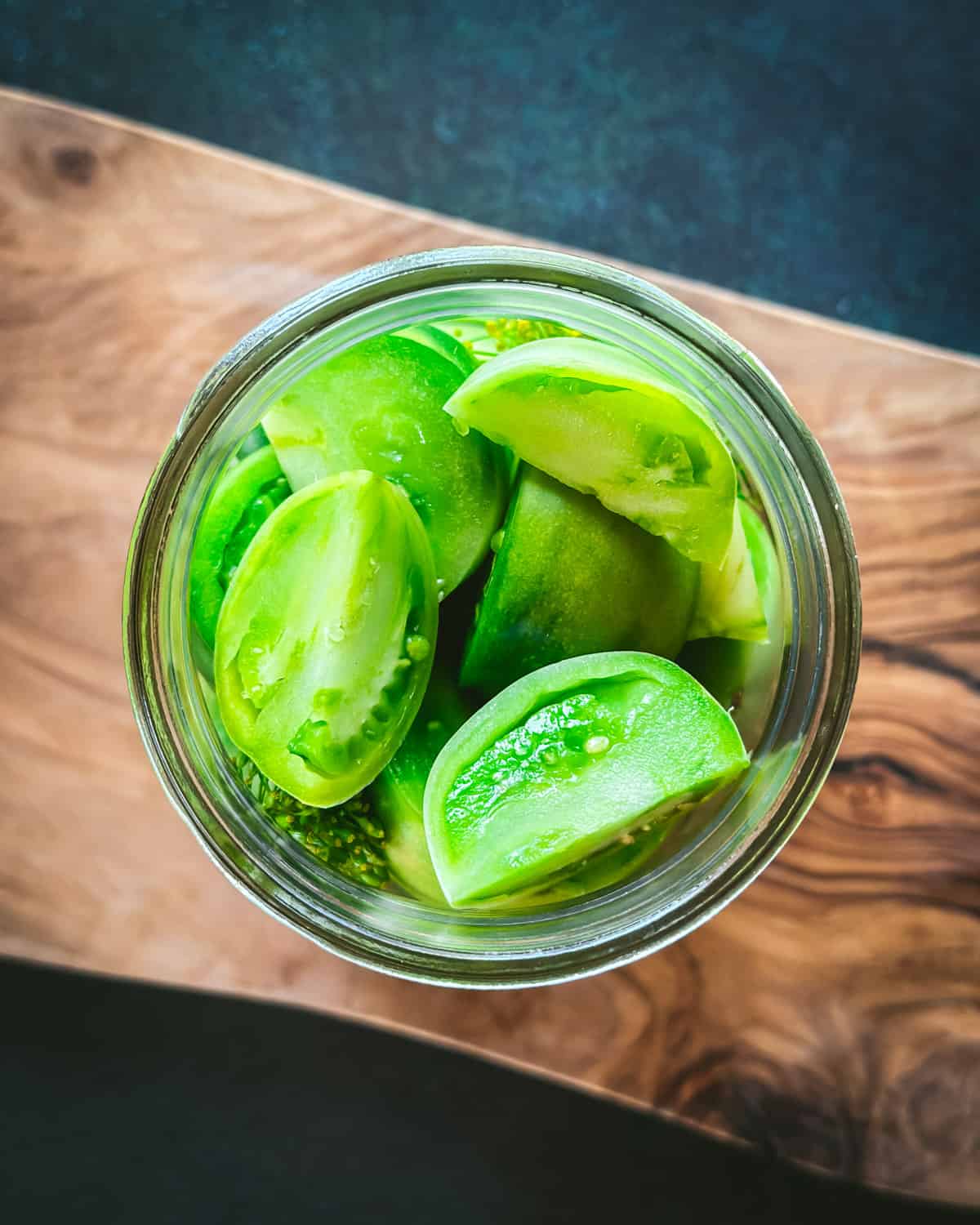 A jar of green tomato wedges on a wood cutting board, ready to be pickled. Top view. 