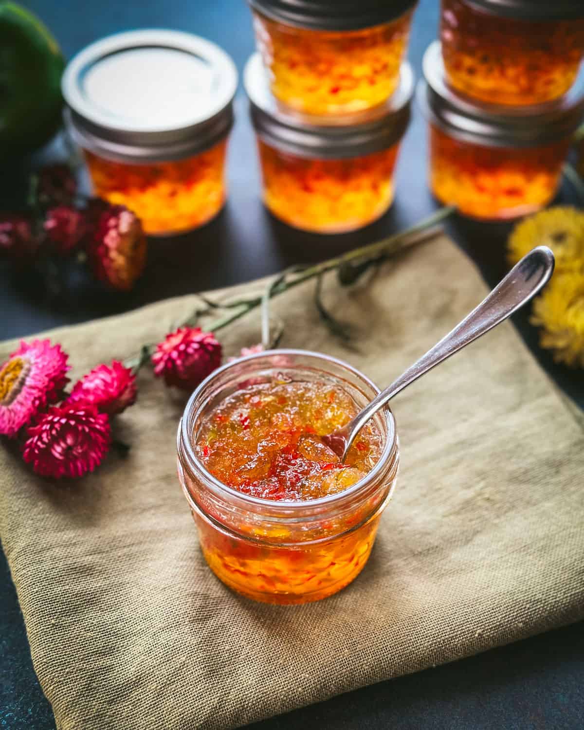 A jar of pepper jelly opened with a spoon resting in it, on a wood cutting board surrounded by flowers and a long homegrown orange bell pepper.