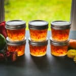 6 small jars of pepper jelly in stacks of 2, with a window in the background letting natural light through the pepper jelly.