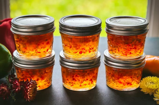 6 small jars of pepper jelly in stacks of 2, with a window in the background letting natural light through the pepper jelly.