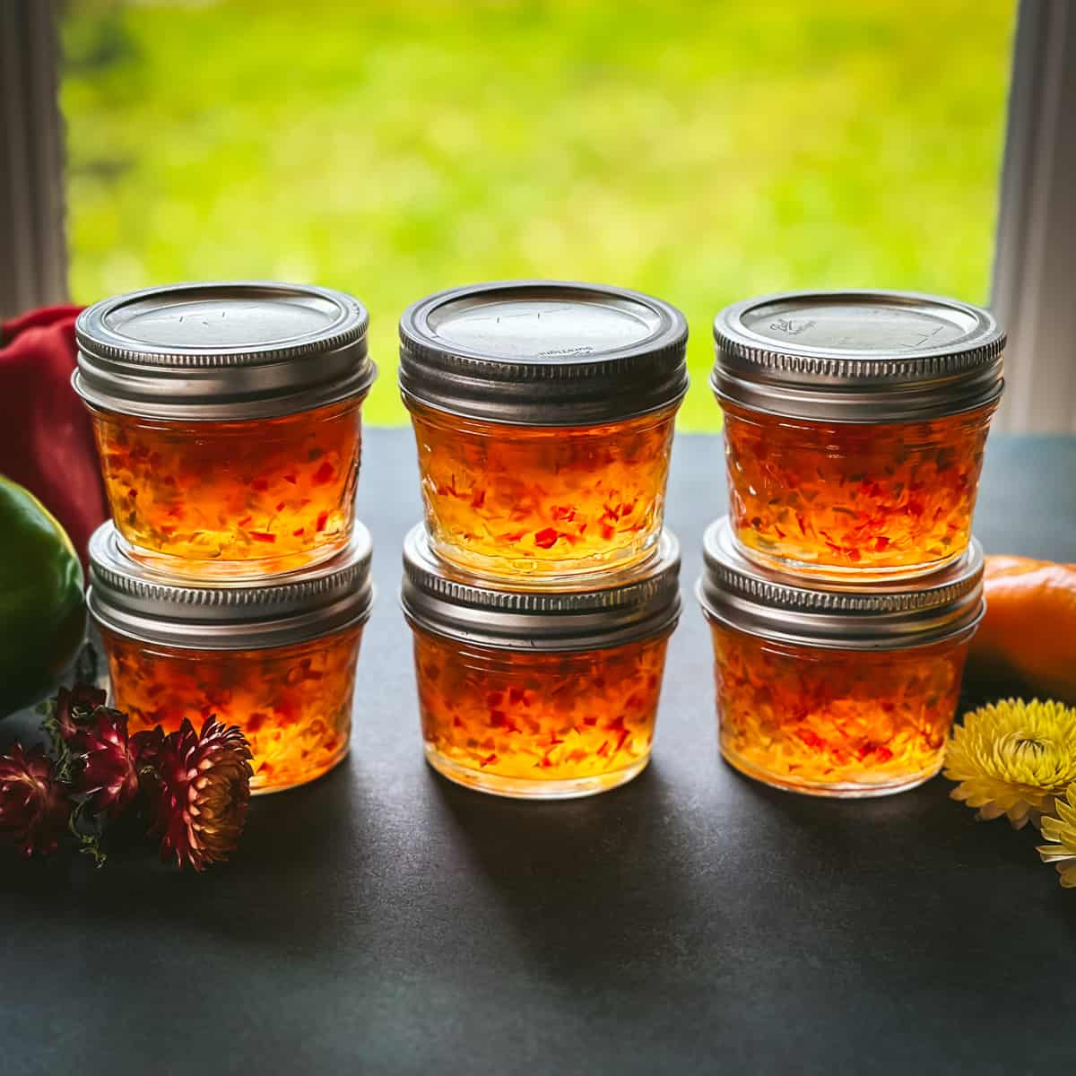 6 small jars of pepper jelly in stacks of 2, with a window in the background letting natural light through the pepper jelly. 